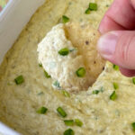 Fingers holding round tortilla chip with artichoke dip and fresh jalapenos on top