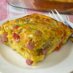 Close up piece of ham, egg and cheese breakfast casserole on a small white plate with a fork in the background