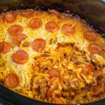 Portion of pizza casserole in crock pot showing noodles, red sauce, cheese and pepperoni in a black crock