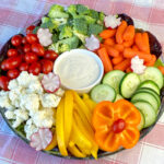 Overhead view of a veggie tray with carrots, cucumbers, bell pepper slices, cauliflower, cherry tomatoes, broccoli and a ranch dressing dip on a red and white plaid napkin