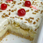 Thick layer of whipped cream on bananas and cream cheese filling sandwiches between a vanilla wafer crust and chopped nuts and cherries on top