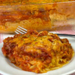 Close up cheesy and saucy plate of delicious Tik Tok alfredo spaghetti with dish in the background showing distinct layers of spaghetti alfredo and red meat sauce on top