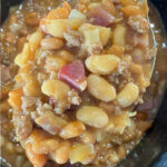 Close up spoon full of meaty baked beans loaded with ground beef and bacon above a crock pot full of warm baked beans