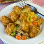 Cheese covered hamburger peeking out of a tater tot casserole with vegetables and a fork leaning on the crispy potatoes in the background