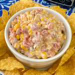 White bowl generously filled with warm mexican corn dip on a bigger plate loaded with tortilla chips on a blue and white napkin