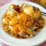 Baked red potato chunks with the skin on coated in ranch dressing, sour cream, cheese and bacon then topped with more cheese and butter soaked cornflakes on a white plate with a fork in the background.