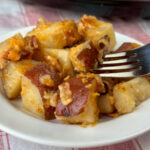 Close up of tender pieces of red potatoes with melted cheese, bacon bits and seasoning coating them and a fork in one piece on a white plate.