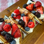 Close up of delicious creamy filled pancake breakfast tacos loaded with fruit and drizzled with chocolate sauce on a wooden table top.