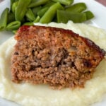 Serving of glazed meatloaf on a pile of mashed potatoes with green beans in the background.