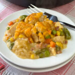 Plate full of chicken and mixed vegetables nestled in gravy and topped with baked golden brown cheesy tater tots with a fork resting in the background.