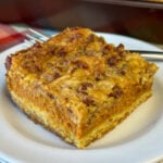 Square piece of delicious pumpkin pie cake with layers of a yellow cake mix, a thick pumpkin layer and a crumbly topping with pecans on a white plate with a fork in the background.