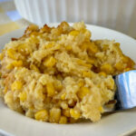 Close up of a fork in a scalloped corn casserole with crackers and cheese on a white plate.
