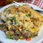 Close up tender ground beef coated with a creamy vegetable, milk and broth mixture with Red Lobster biscuit mix baked in a pot pie crust layer on top on a red and white plaid napkin.