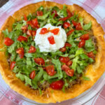 Close up of a delicious taco pie topped with lettuce, tomatoes and sour cream on a red and white plaid napkin.