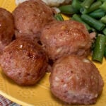 Close up of ham balls with a sweet and smoky flavor and a light golden brown finish on a yellow plate with green beans and mashed potatoes in the background.