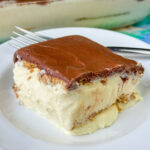 Chocolate frosting covering a graham cracker resting on a creamy vanilla filling with more layers of graham crackers on a white plate with a fork in the background.