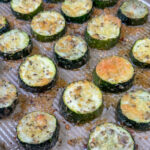 Parmesan cheese melted on round slices of roasted zucchini on a ribbed baking sheet.