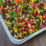 An economical side dish salsa made with black beans and corn creating earthy flavors that are complimented with fresh tomatoes, green onions and cilantro and coated with a light lemon and olive oil dressing in an aluminum pan.