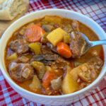 Close up bowl full of tender stew meat in a hearty tomato based sauce that is loaded with potatoes, carrots, onion, celery and mushrooms to create our amazing Crock Pot Beef Stew Recipe on a red and white napkin.