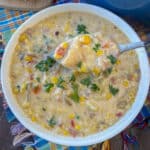 Spoon lifting a bite of corn chowder with bacon and potatoes above a bowl full of flavorful soup.