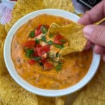 Hand scooping cheeseburger dip garnished with chopped tomatoes and parsley on a tortilla chip in a small white dish.