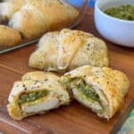 Warm, flaky crescent roll baked with a filling of pesto over caprese cheese cut in half with more rolls in the background on a wooden board.