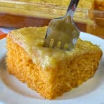 Square piece of orange cake with a layer of pineapple frosting and a fork in the top.