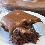 Close up moist piece of old fashioned chocolate cake with chocolate frosting on a fork in front of the square piece of cake.