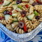 Close up corner of a foil pan filled with twist pasta coated with Balsamic Vinaigrette dressing with olives, cucumbers and roasted red peppers.