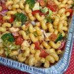 Corner of a foil pan filled with seasoned twist pasta surrounded by broccoli, cauliflower, tomatoes, pepperoni and cheese.