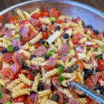 Close up chopped salami in pasta salad with Italian dressing, cheese, olives, tomatoes and green peppers.