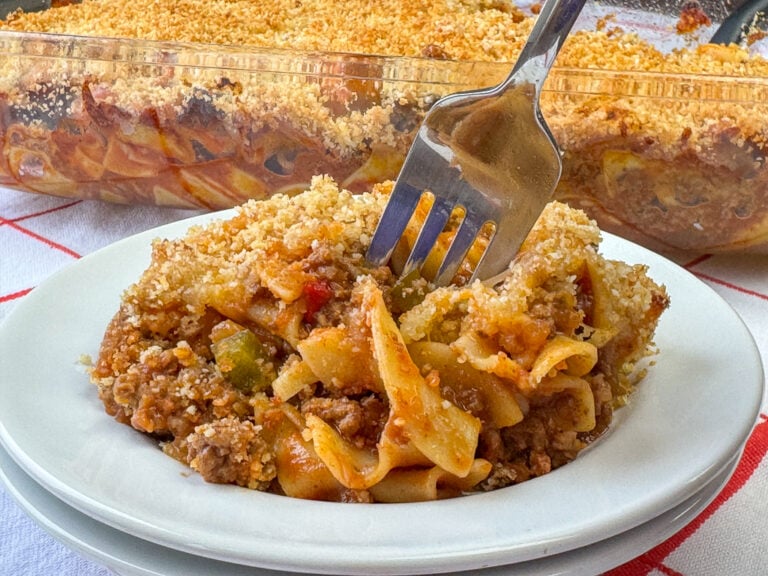 Plate and fork in a serving of a tasty Mexican casserole with ground beef and egg noodles with an easy sauce and a few vegetables in front of a full casserole in the background.