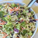 Broccoli slaw with pieces of apple and pineapple stirred in then coated with a simple poppyseed dressing in a white bowl with a serving spoon on a yellow and white plaid napkin.