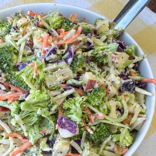Broccoli slaw with pieces of apple and pineapple stirred in then coated with a simple poppyseed dressing in a white bowl with a serving spoon on a yellow and white plaid napkin.