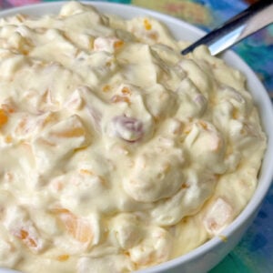 Close up cherry, pineapple tidbit, grape and other fruit cocktail ingredients coated with creamy vanilla pudding mixture heaped up in a white bowl.