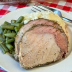 Close up of tender slices of properly cooked pork loin on a plate of fluffy mashed potatoes and cut green beans with a fork in the background.