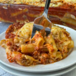 Serving of smooth tubes of ziti pasta covered in a tomato based meat sauce and melted cheese with a fork in the center on a white plate.