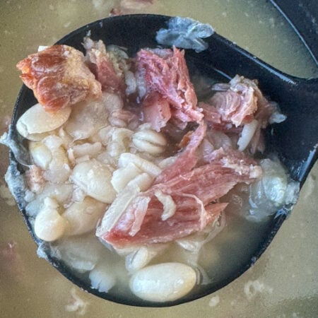 Soaked northern white beans in a flavorful broth with shredded bits of ham in a soup ladle.