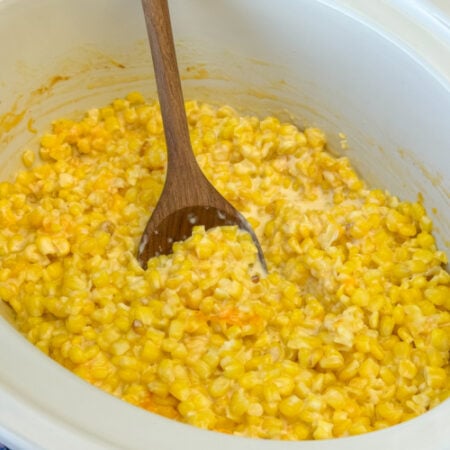 Frozen corn coated with cheddar cheese in a cream cheese sauce with a wooden spoon stirring.