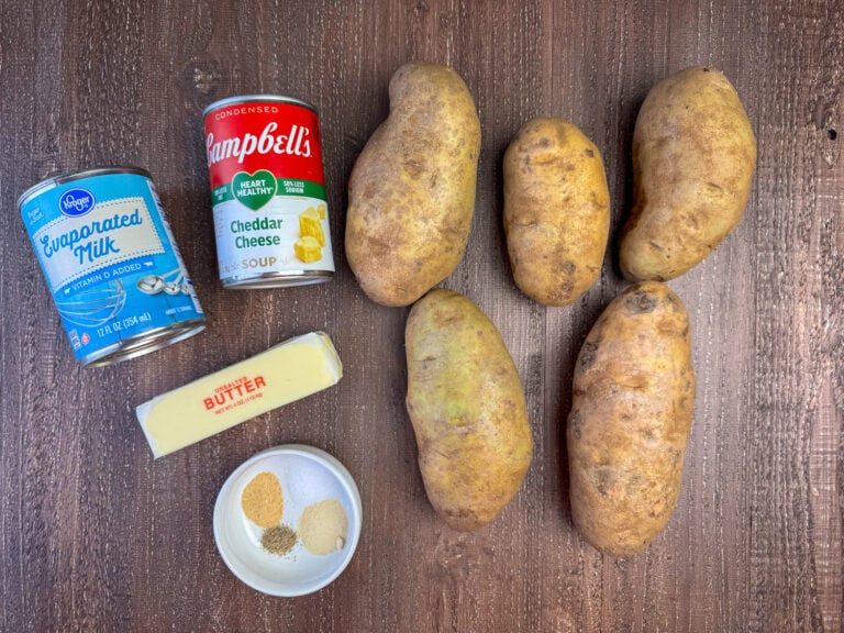Ingredients to make crock pot potatoes are evaporated milk, cheddar cheese soup, large potatoes, butter, salt, pepper, onion powder and garlic powder on a wooden counter top.