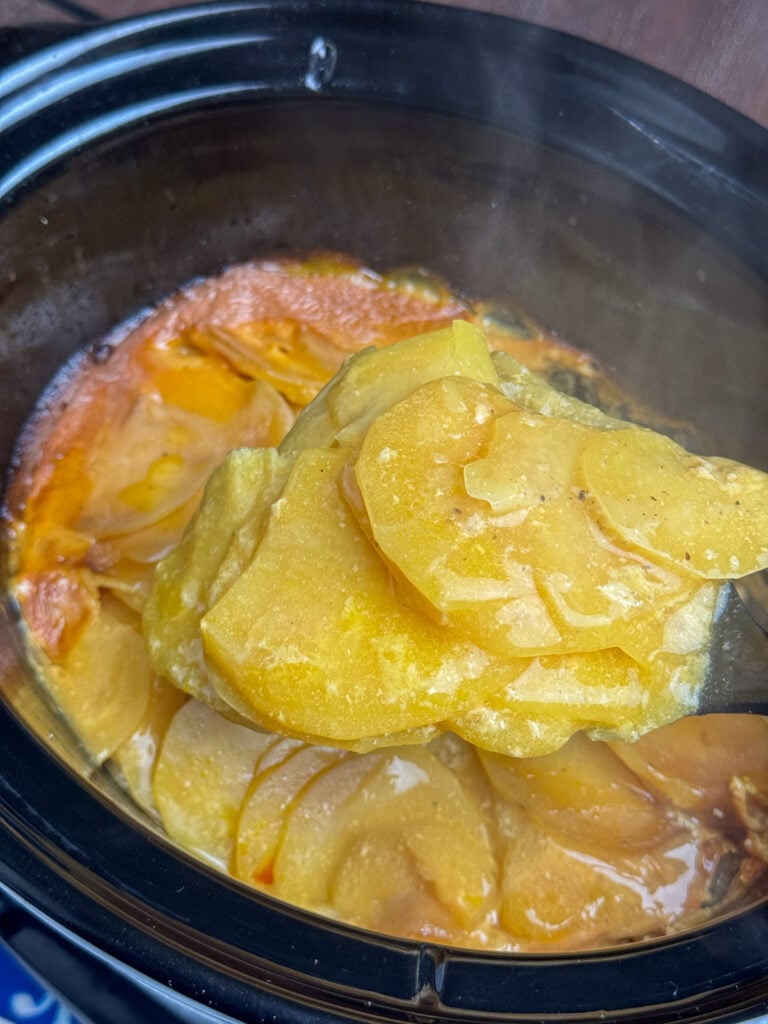 Spoon lifting a serving of tender and slightly cheesy slices of potatoes out of a hot crock pot.