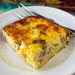 Square piece of a delicious potato, egg, ham and cheese casserole on a white plate with a fork leaning on the edge.