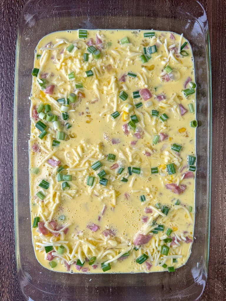 Scrambled eggs poured on top of layers of potatoes, ham, cheese and green onion in a glass dish on a wooden counter top.