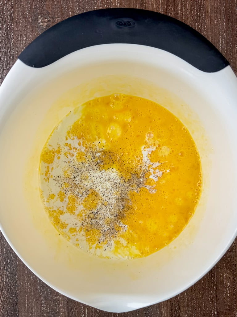 Eggs and evaporated milk seasoned with salt and pepper cracked into a white mixing bowl.