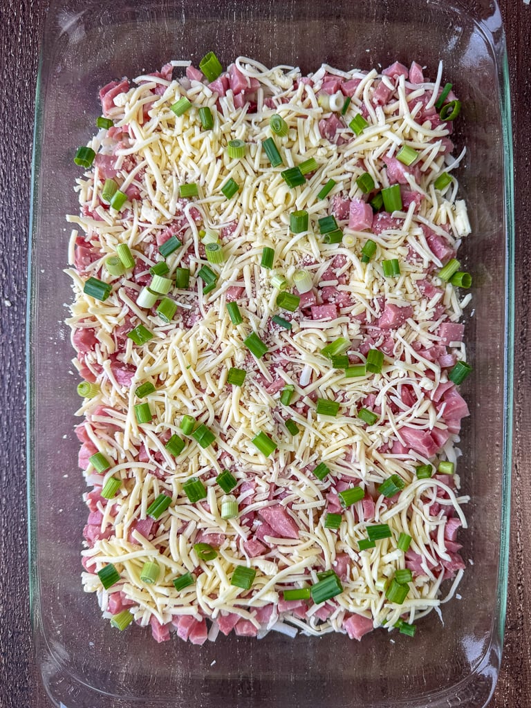 Chopped green onions sprinkled on shredded cheese, cubes of ham and a layer of potatoes in a glass 9 x 13" casserole dish.