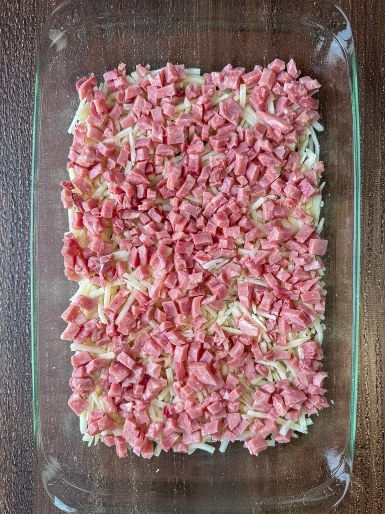 Thick layer of diced ham on top of shredded potatoes in a glass baking dish.