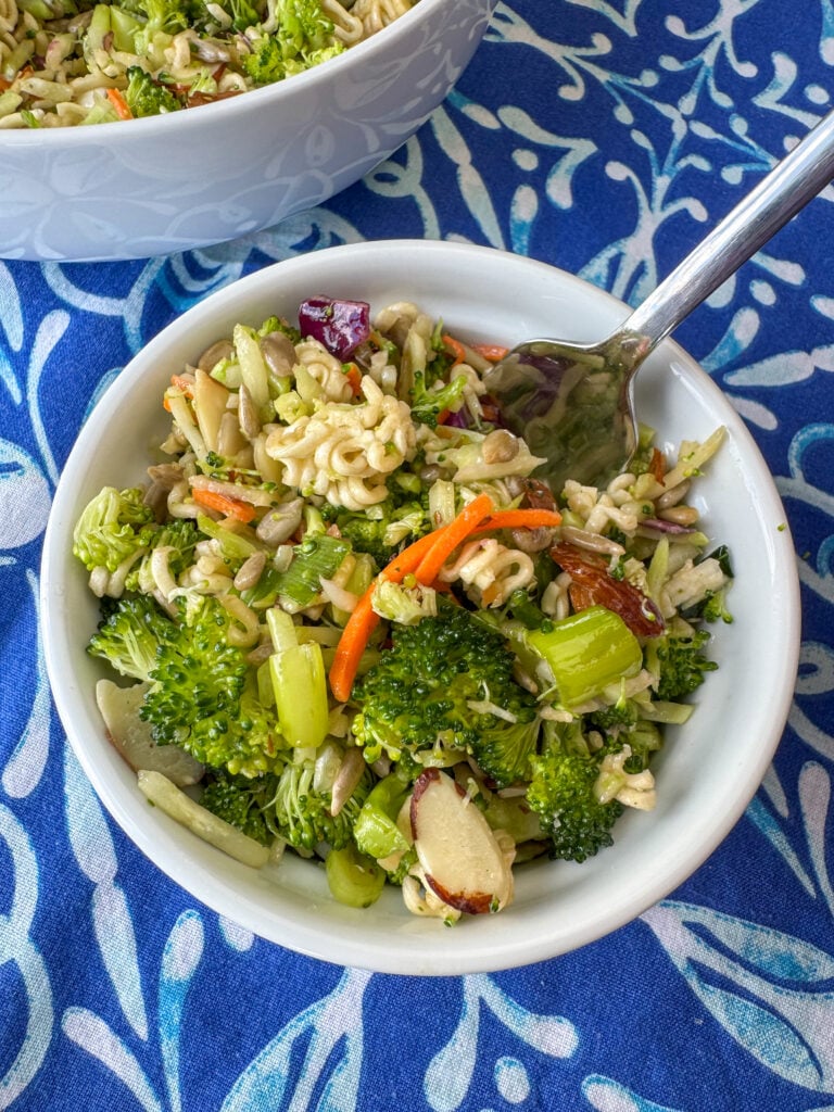 Firm and crunchy broccoli salad enhanced with a simple Asian dressing and ramen noodles in a small white dish next to a bigger bowl full on a blue napkin.