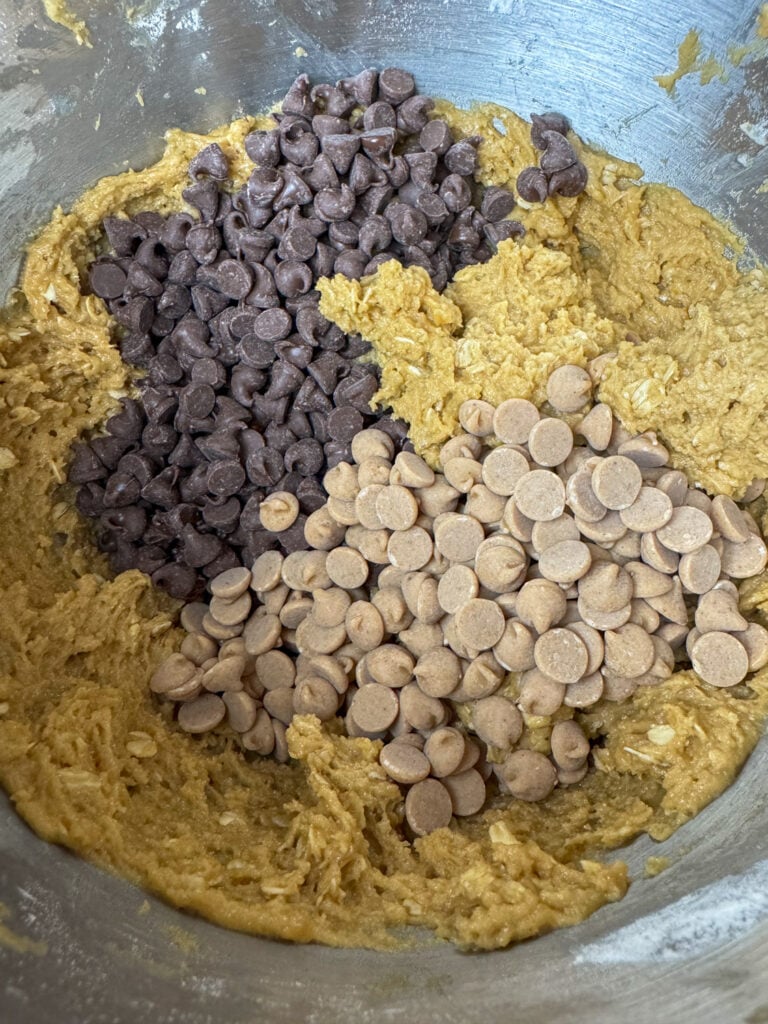 Semi sweet chocolate chips and peanut butter chips poured on top of an oatmeal and peanut butter cookie dough in a silver mixing bowl.