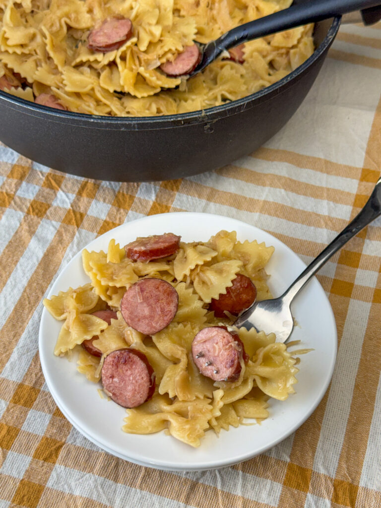 Serving spoon in a pot of Kielbasa pasta and sauerkraut with a serving on a white plate and a fork ready to eat on a yellow and cream napkin.