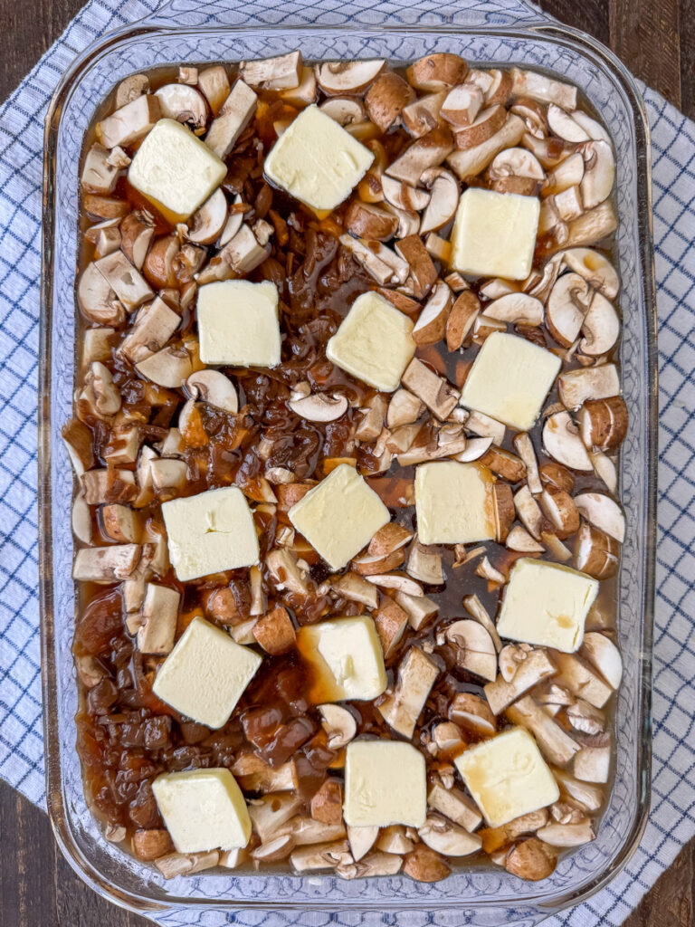 Slices of butter spread over top of chopped mushrooms, French onion soup, beef consomme, rice and ground beef in a glass dish on a blue and white napkin.